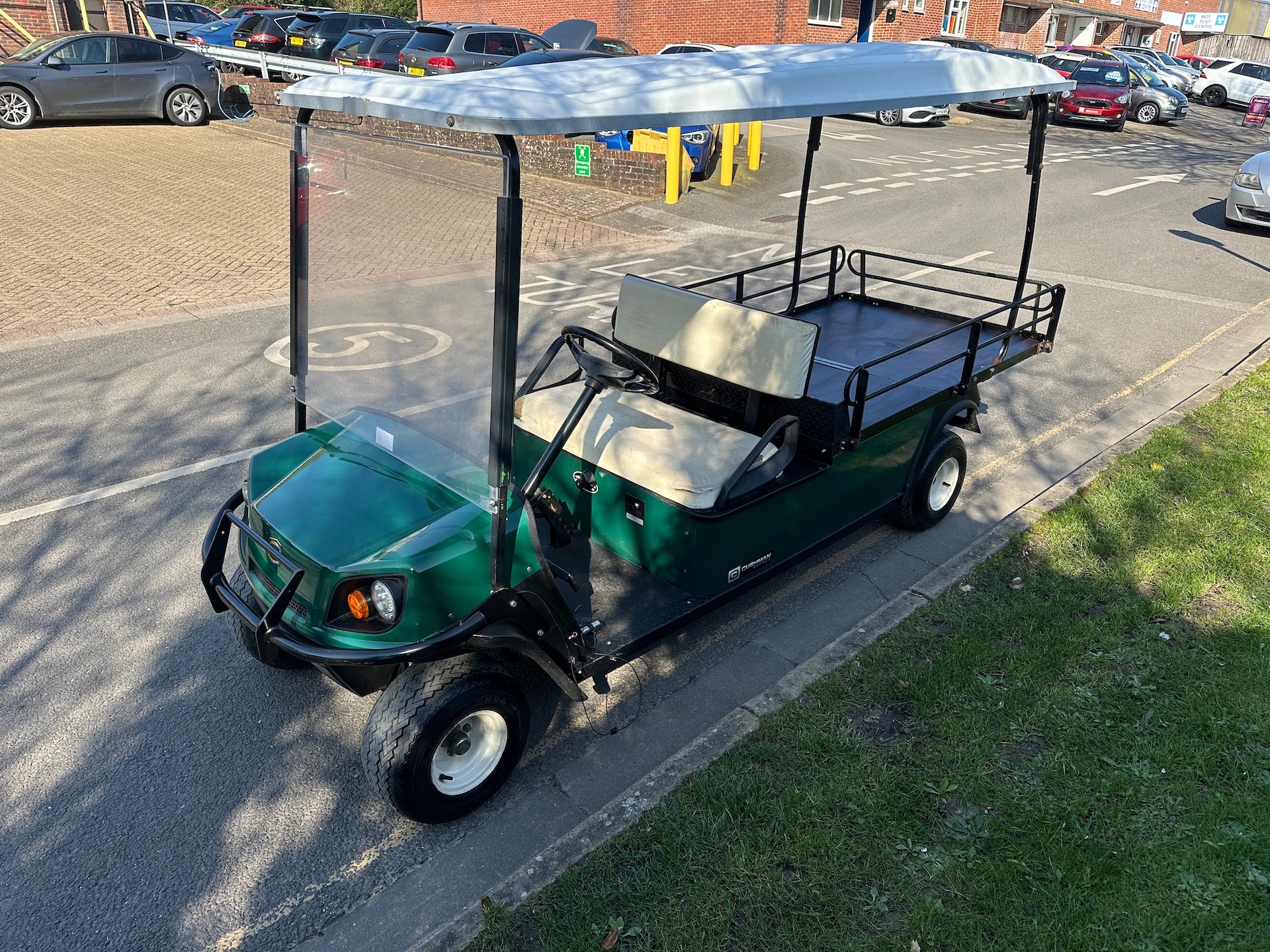 A green four-seater golf cart with a white canopy and black frame, parked on a paved street next to a grassy area in an outdoor parking lot.